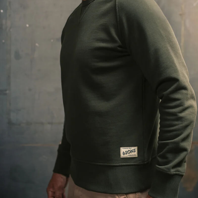Person wearing a green sweater with a brand logo against a grey wall
