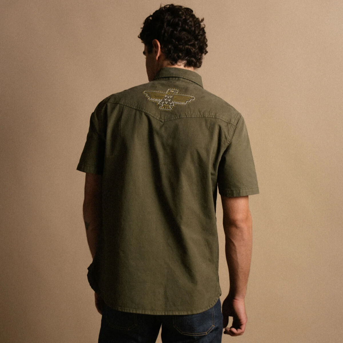Man wearing an olive green shirt with a logo on the back against a beige background
