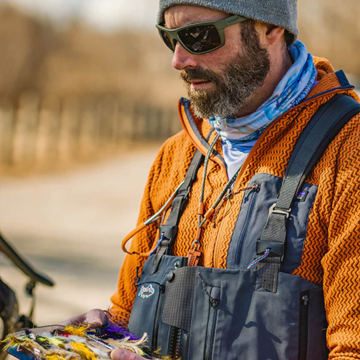 Man in outdoor gear holding fishing lures with a blurred natural background