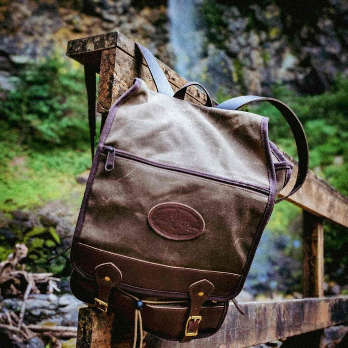A brown waxed canvas backpack with dark brown leather trim and a circular embossed logo patch, resting on a rustic wooden railing with a blurred outdoor waterfall and greenery background.