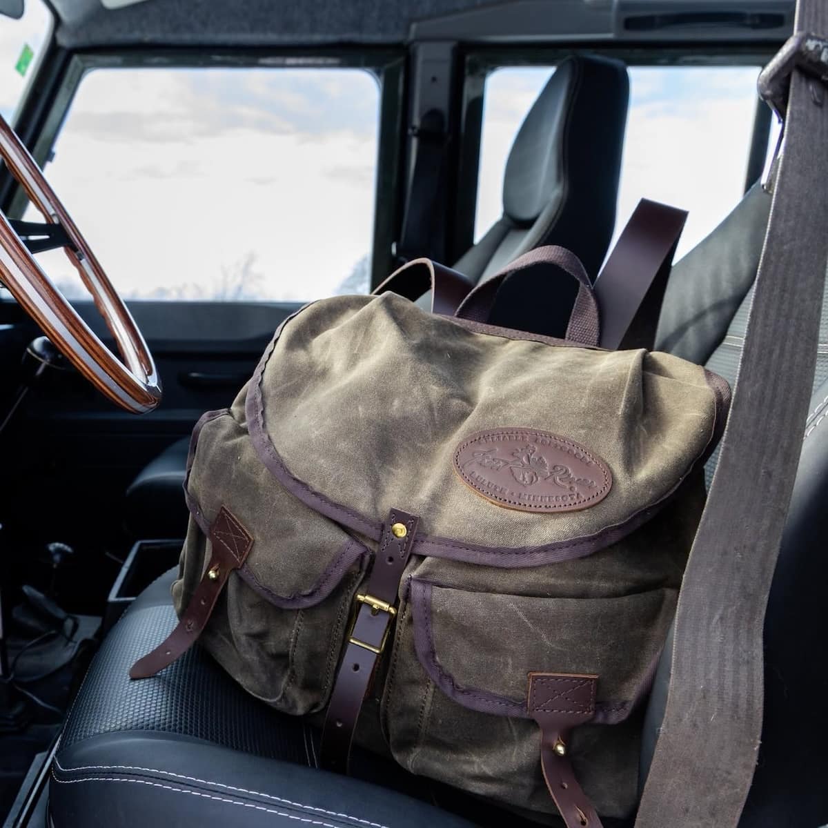 A vintage-style waxed canvas backpack in olive green with dark brown leather straps and trim, resting on a car seat with the driver's seat and steering wheel visible in the background.