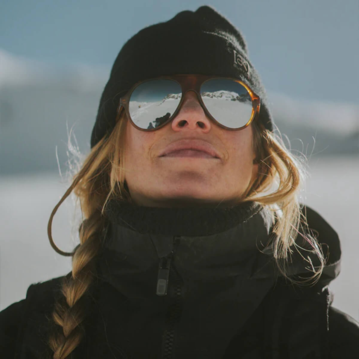 Person wearing sunglasses and a black beanie with a snowy landscape in the background