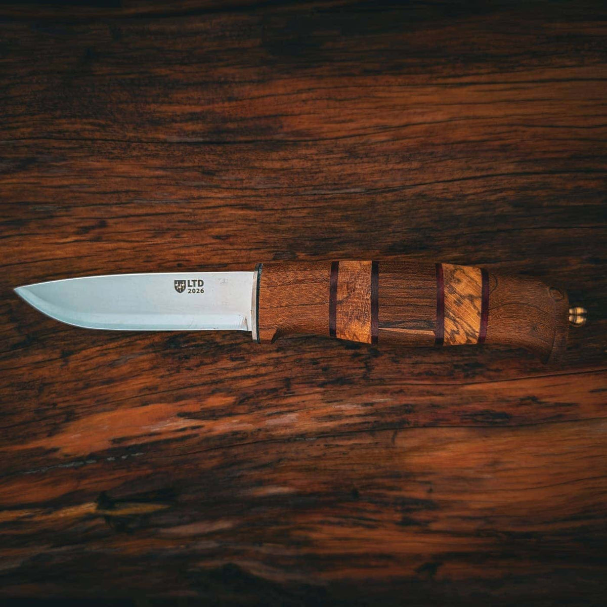 A fixed-blade knife with a silver steel blade and a brown wood-grain handle, displayed horizontally on a dark wood surface. The blade is engraved with the brand name Helle