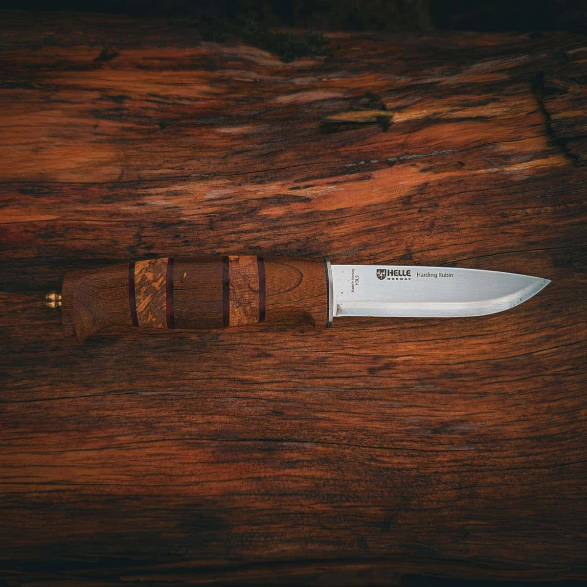 A fixed-blade knife with a silver steel blade and a brown wooden handle with inlaid details, resting on a dark, textured wooden surface. The brand 'Helle' is visible on the blade.