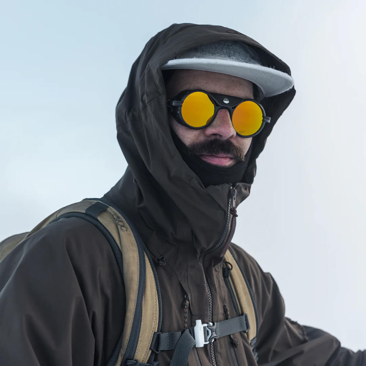 Person wearing a dark hooded jacket and sunglasses with a light background