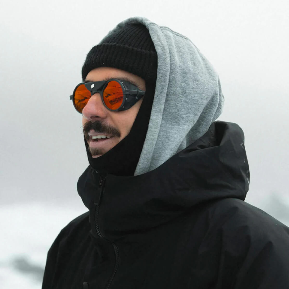Person wearing a black jacket, gray hood, and reflective sunglasses against a blurred background