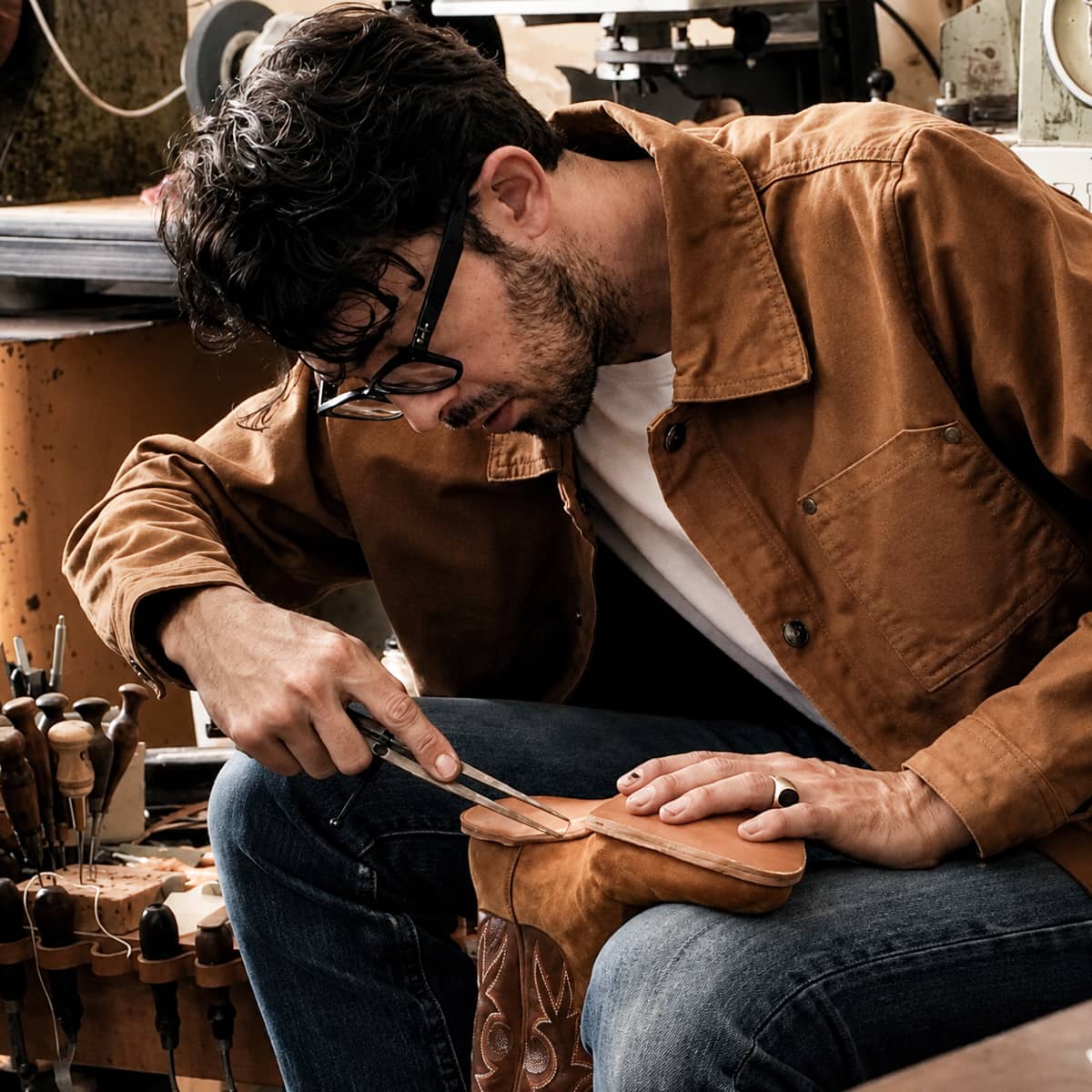 A man wearing an Iron and Resin Jacket sits in a workshop, carefully crafting a brown leather boot on a workbench.