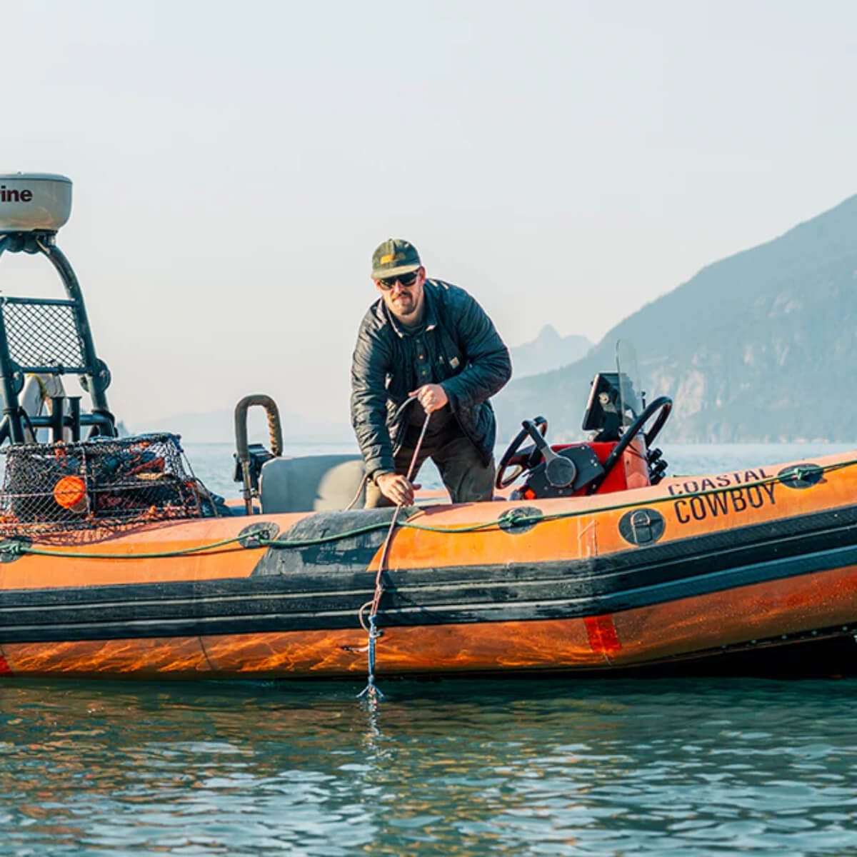 Man on an orange inflatable boat with 'Coastal Cowboy' branding, mountains in the background.
