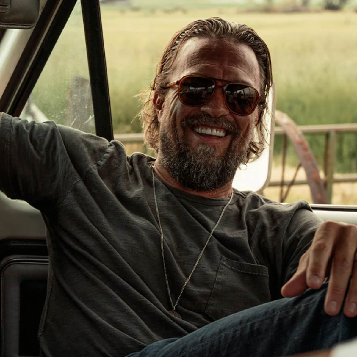 Man with sunglasses and a beard sitting inside a vehicle with a natural landscape outside.