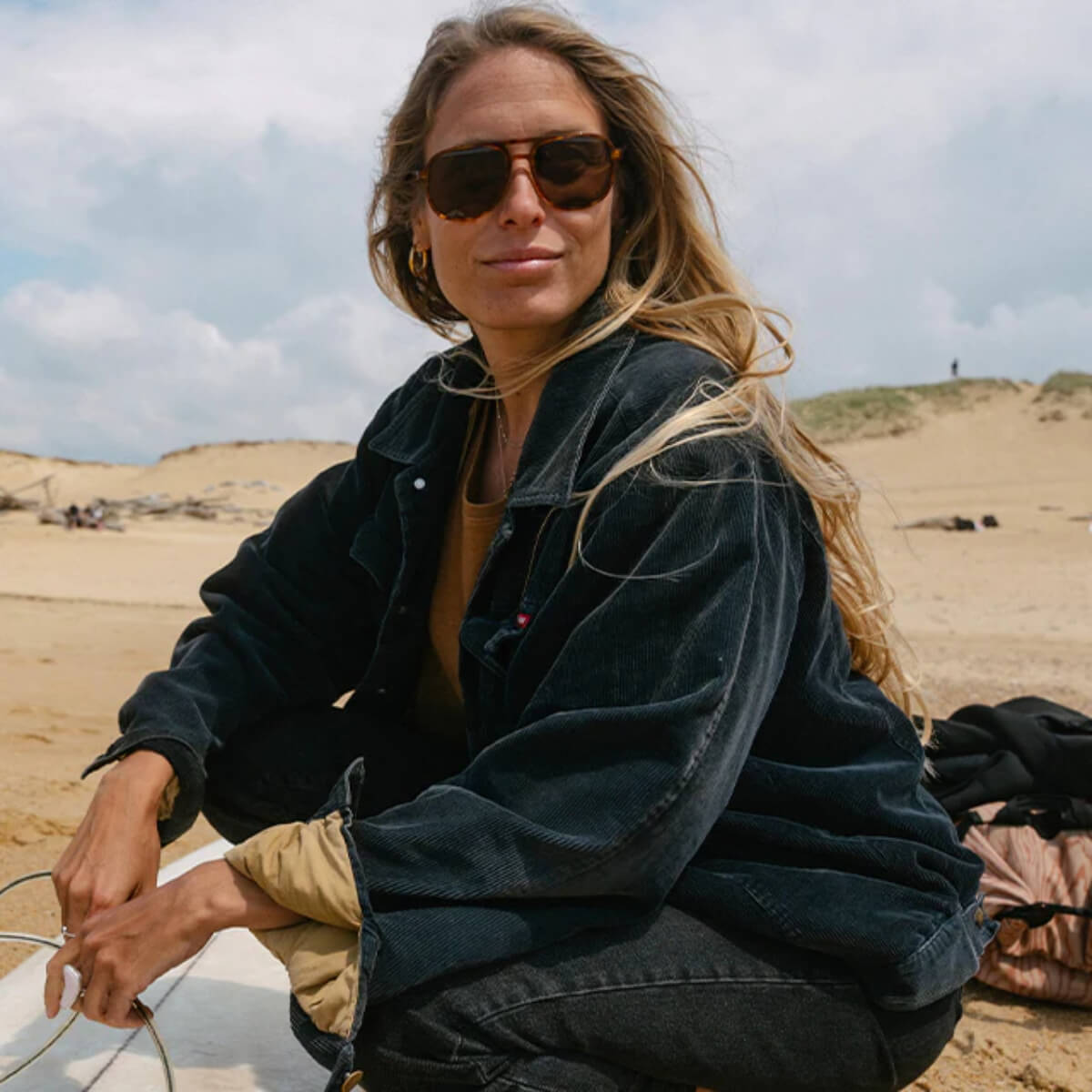 Woman in a black jacket and sunglasses sitting on a sand dune.