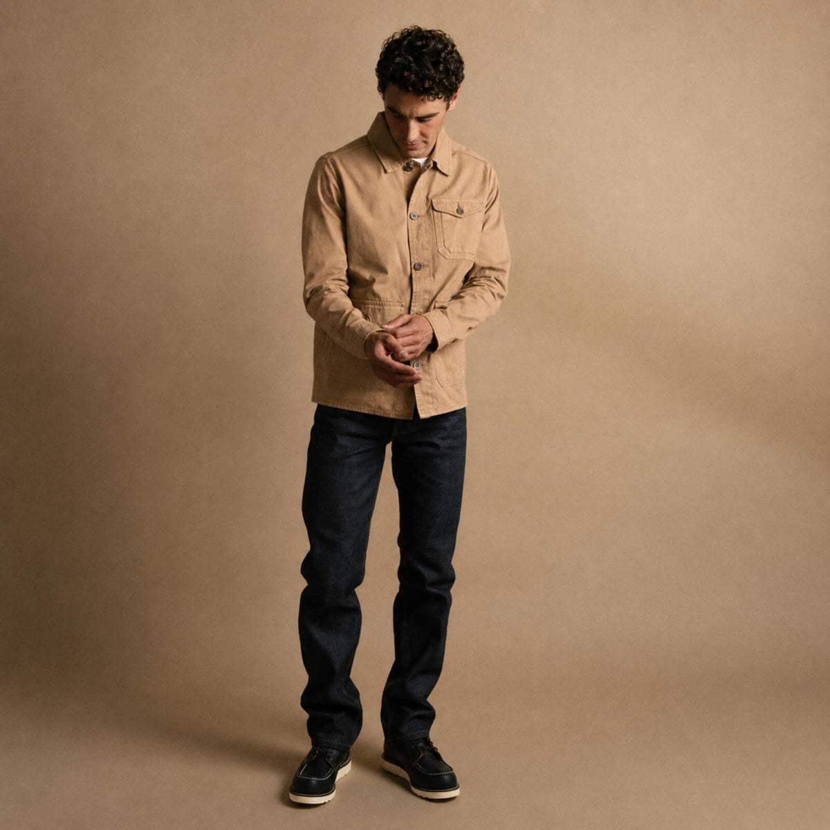 Man wearing a beige jacket and dark jeans against a brown background
