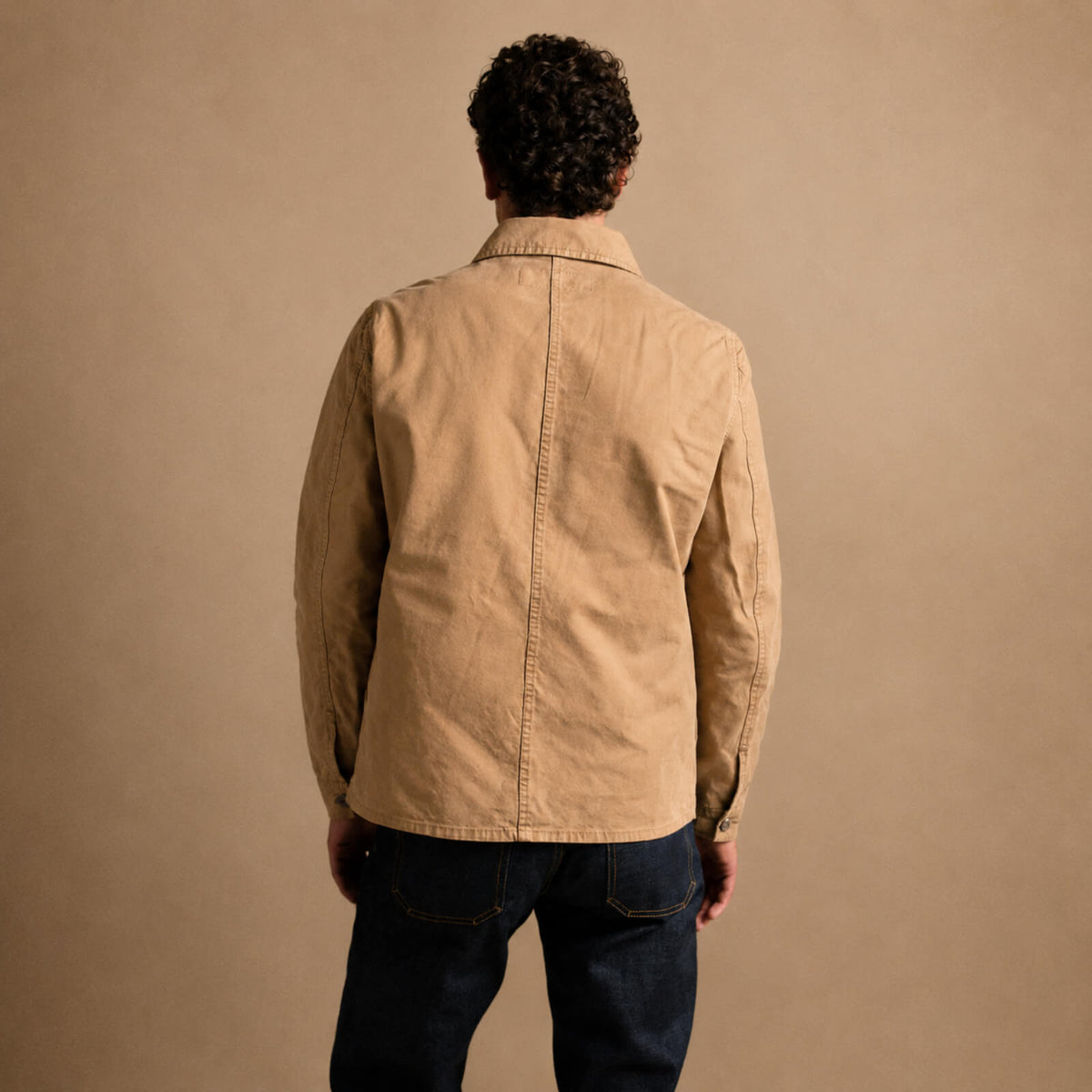 Person wearing a beige jacket and dark jeans against a brown background