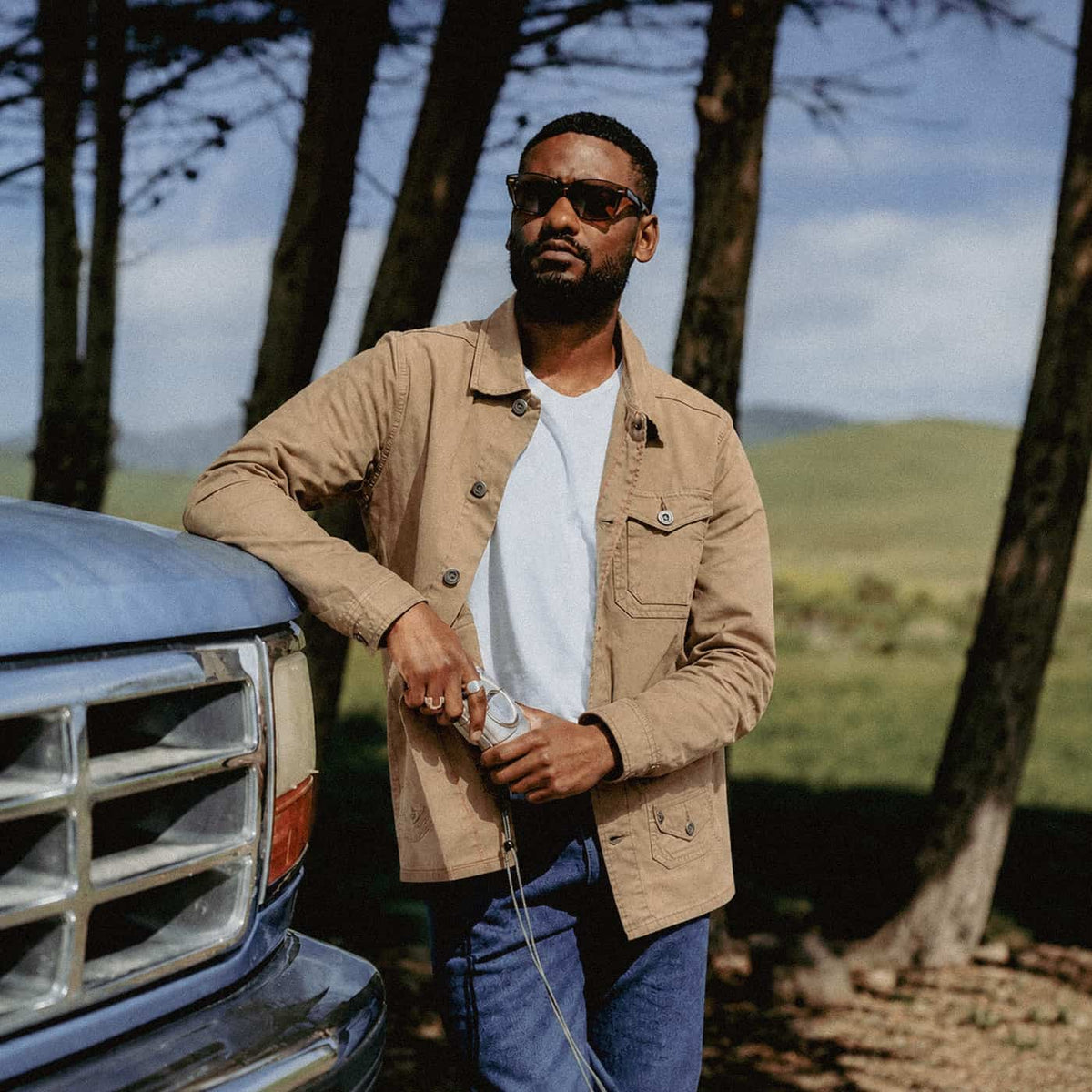 A man wearing a tan utility jacket over a white t-shirt and blue jeans stands outdoors next to a blue truck, holding a camera. Trees and a grassy field are visible in the background.