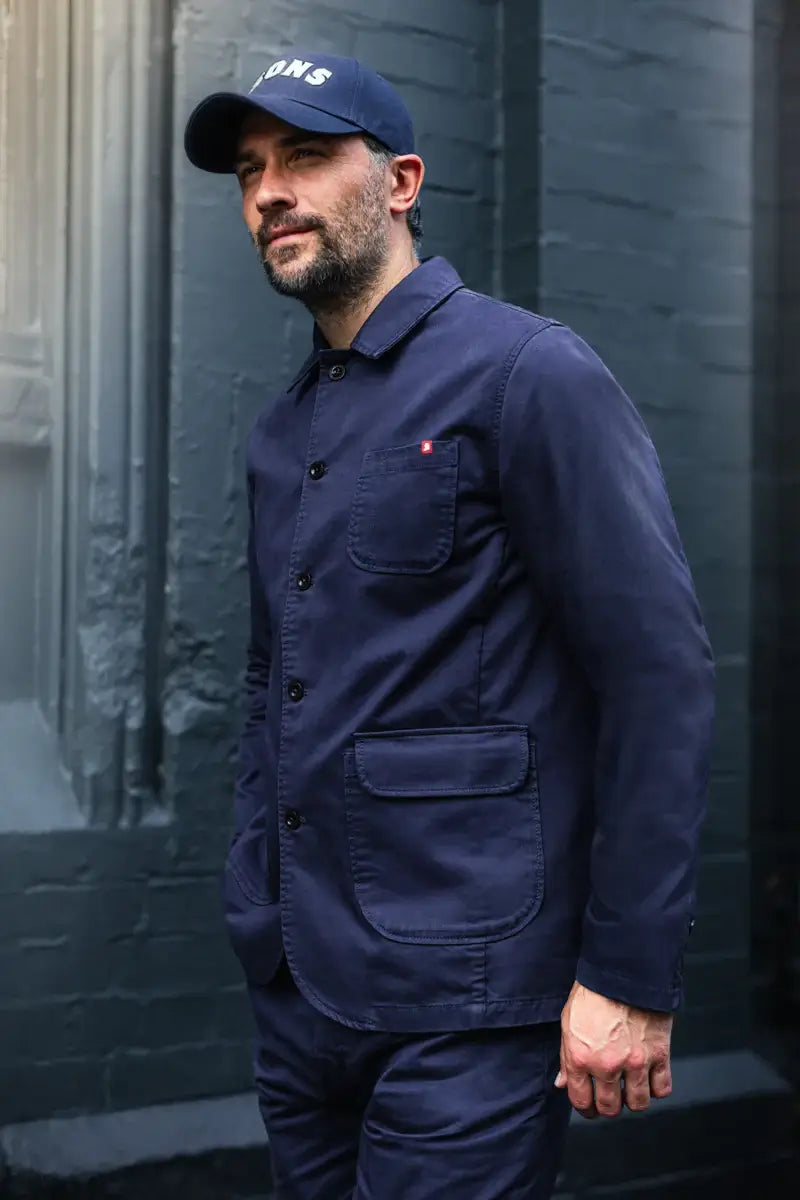 Man wearing a navy blue jacket and cap standing against a dark brick wall.