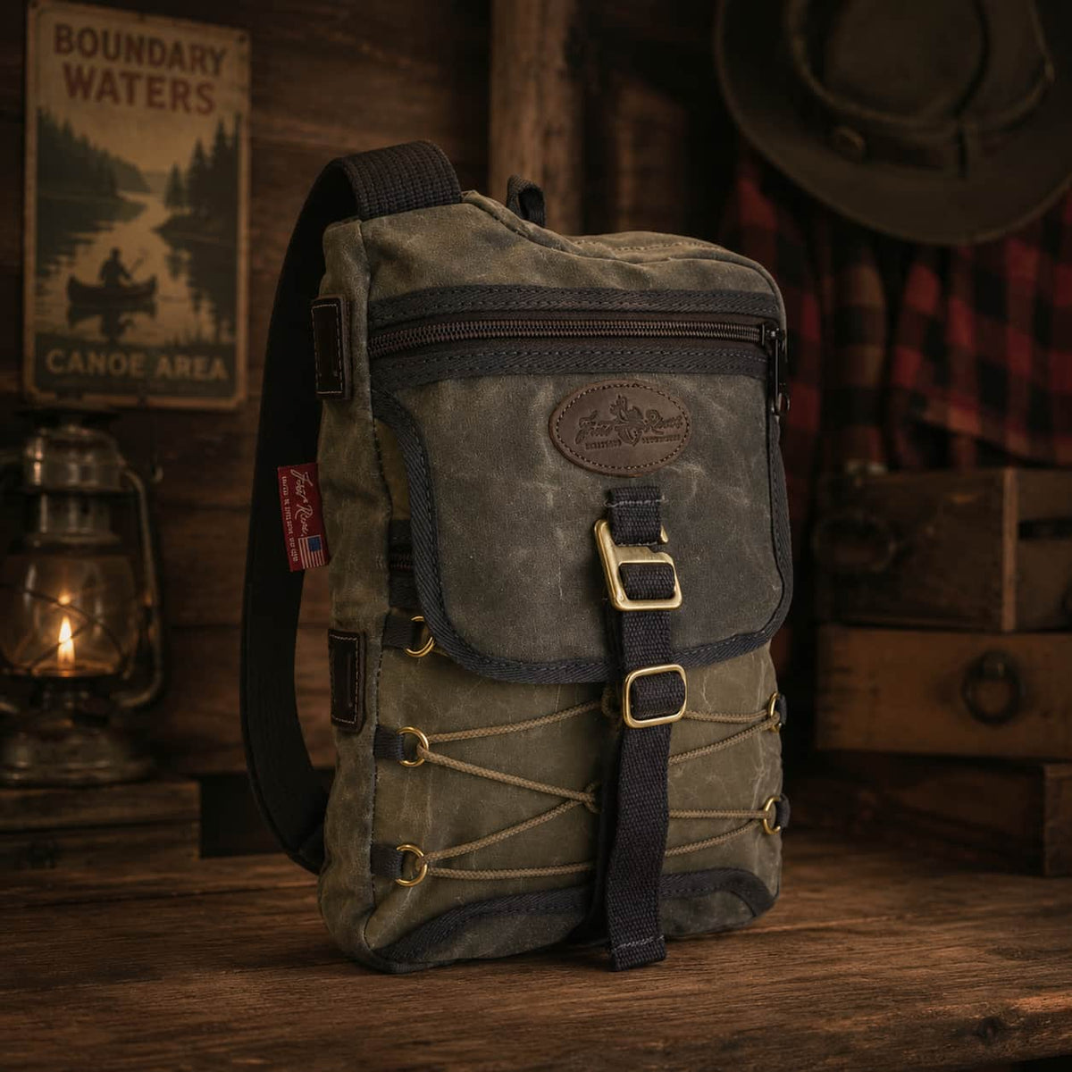 A rugged canvas crossbody bag in distressed olive green with dark navy trim and brass-tone hardware, standing on a wooden surface in a rustic cabin-style setting with a lantern, sign, hat, and plaid shirt in the background.