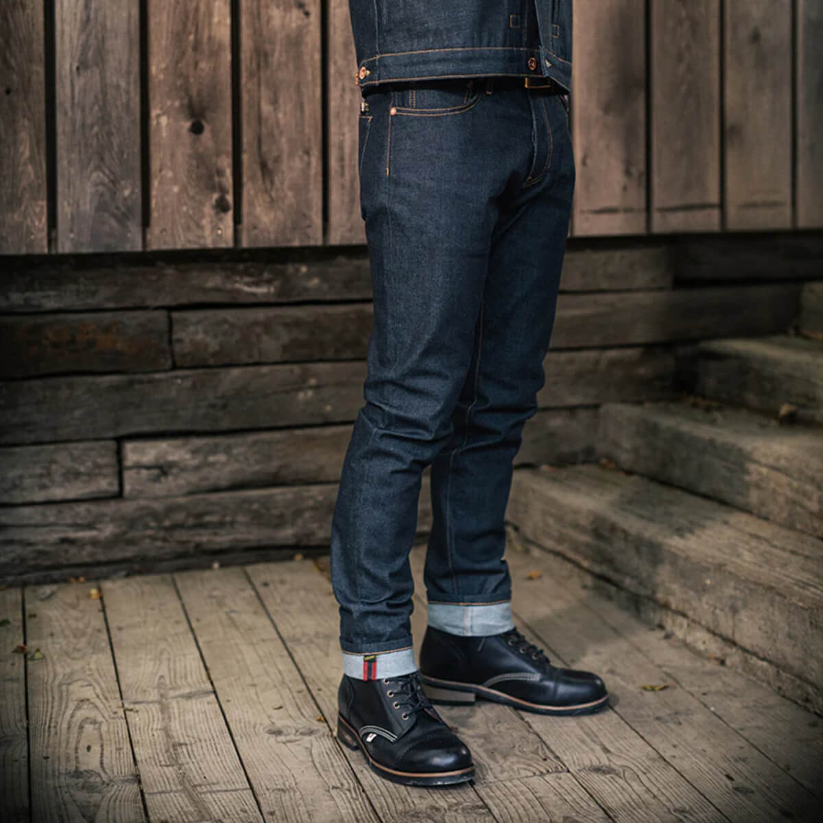 Person wearing dark blue jeans and black shoes standing on wooden steps with a rustic wooden wall background.