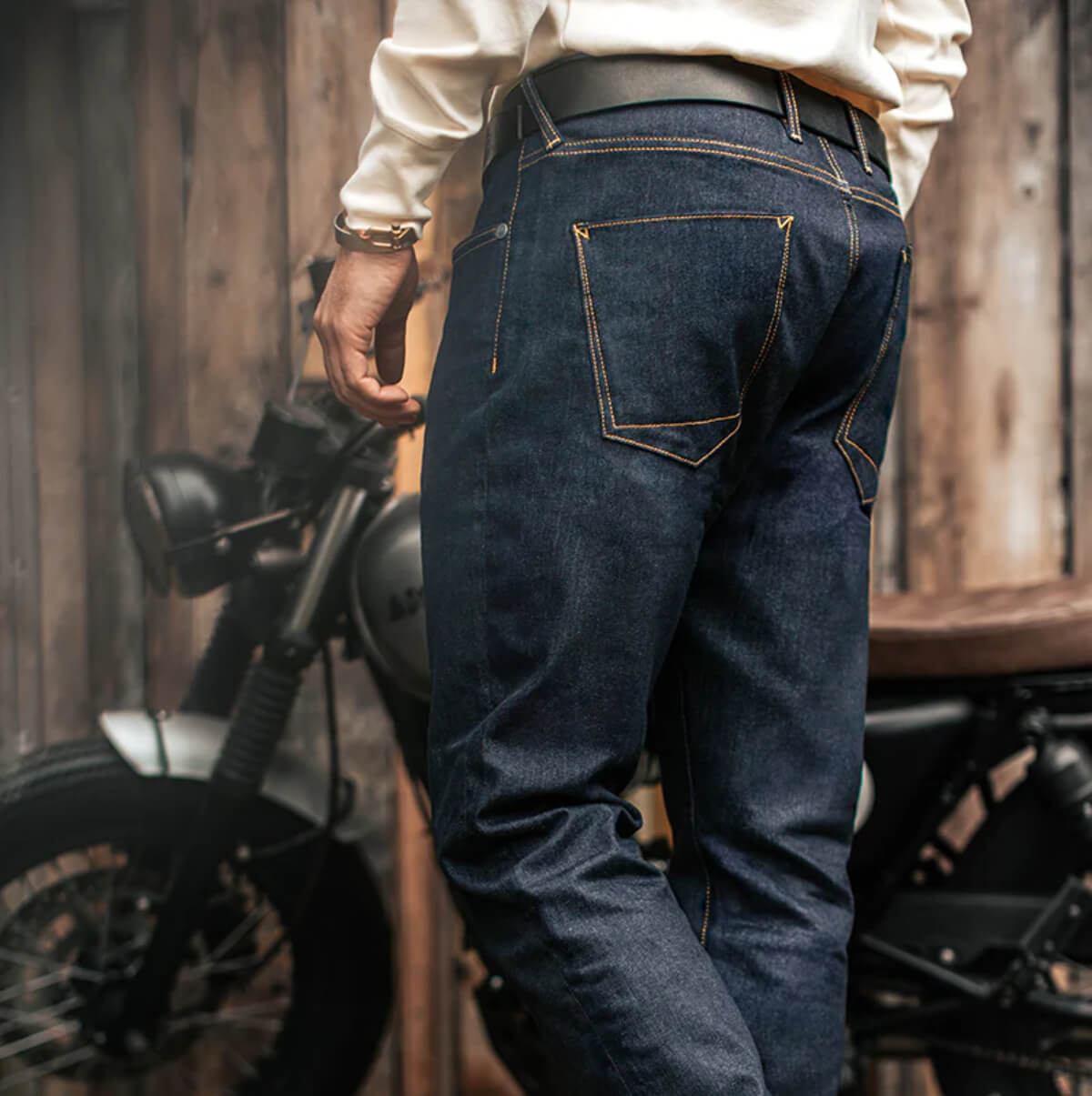 Person wearing dark blue jeans standing next to a motorcycle in a wooden setting
