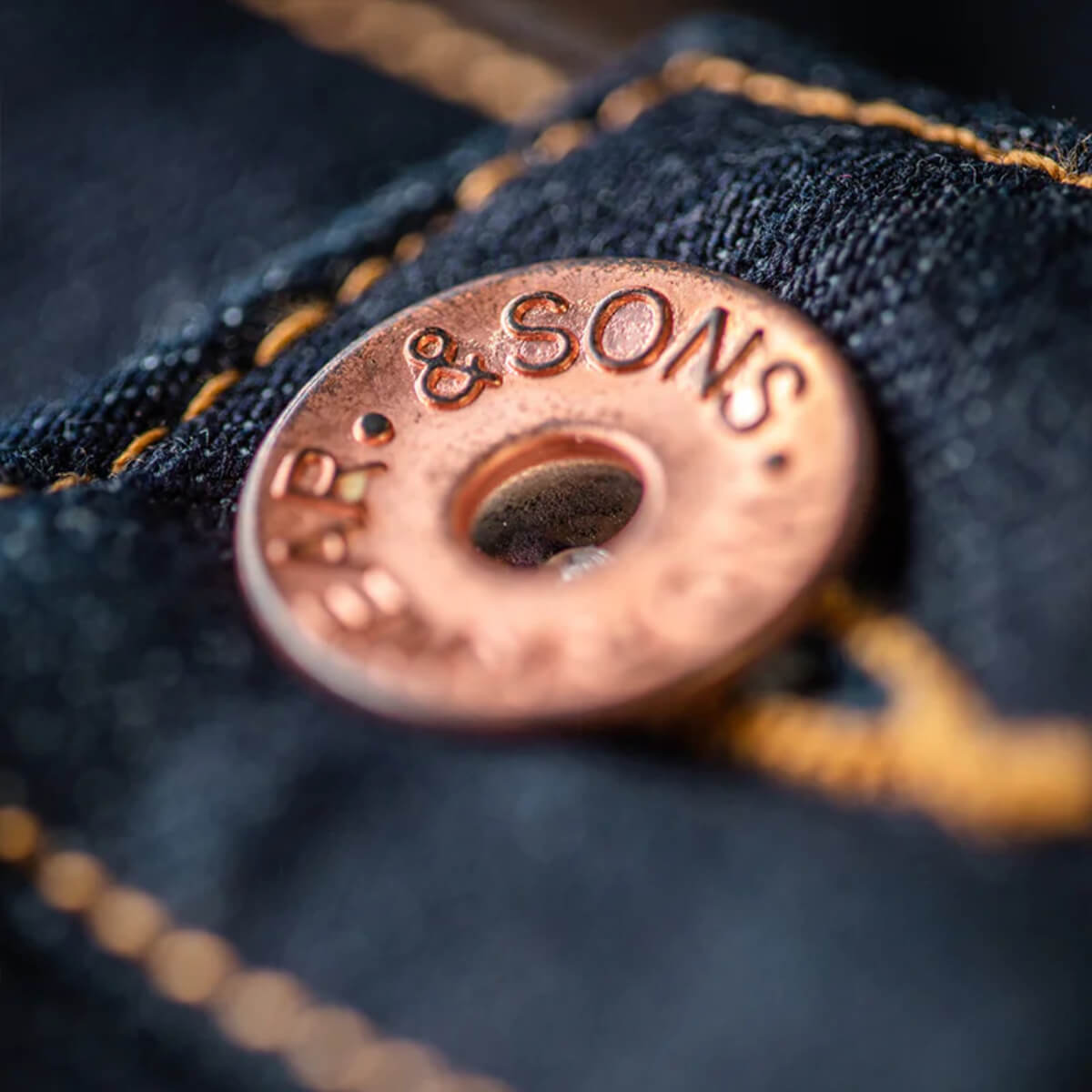 Close-up of a button on a denim fabric with 'R. & SONS' branding.