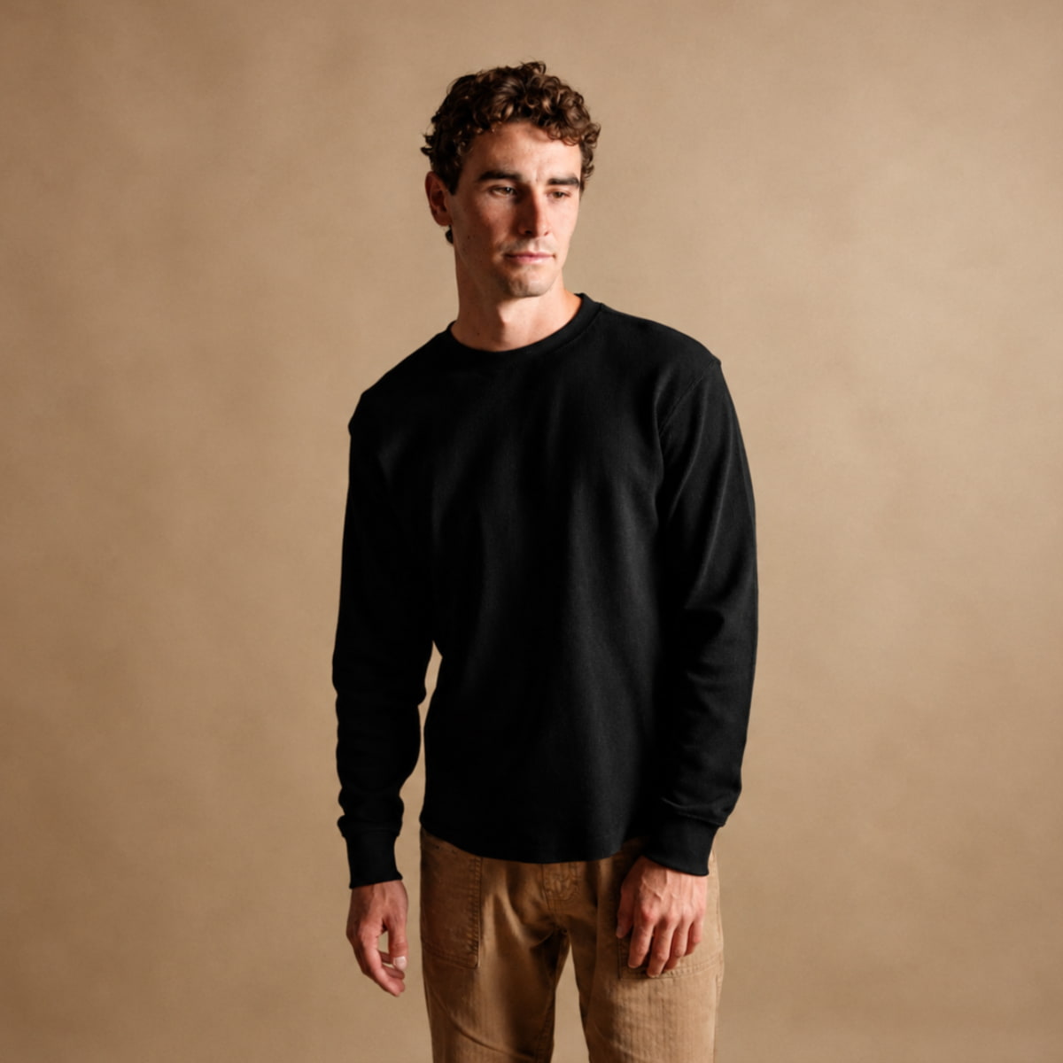 A man wearing a plain black Iron and Resin Cotton Crewe Neck and tan pants, standing against a solid warm beige studio background.