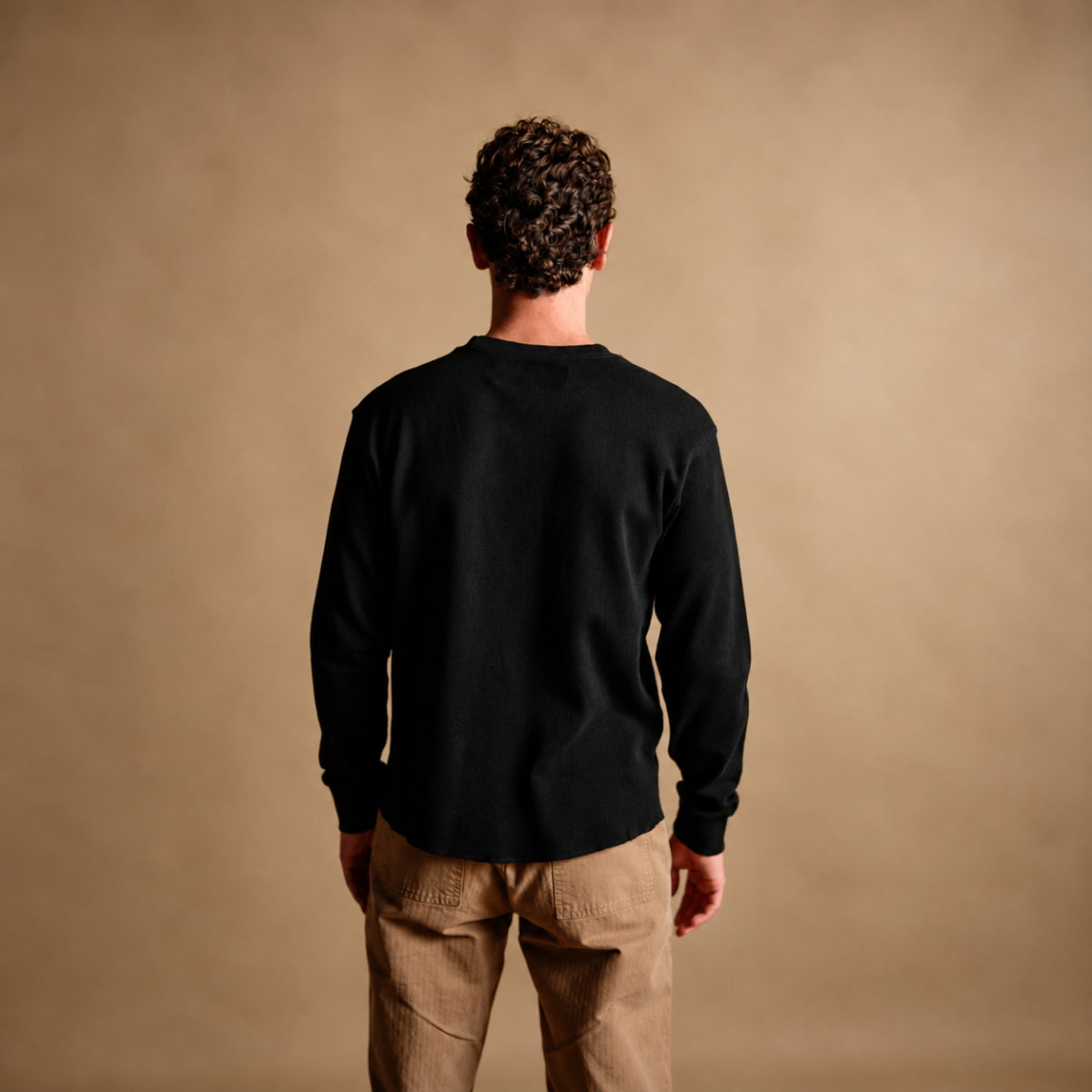 A man viewed from behind wearing a black Iron and Resin Cotton Crewe Neck and tan pants, standing against a solid warm beige studio background.