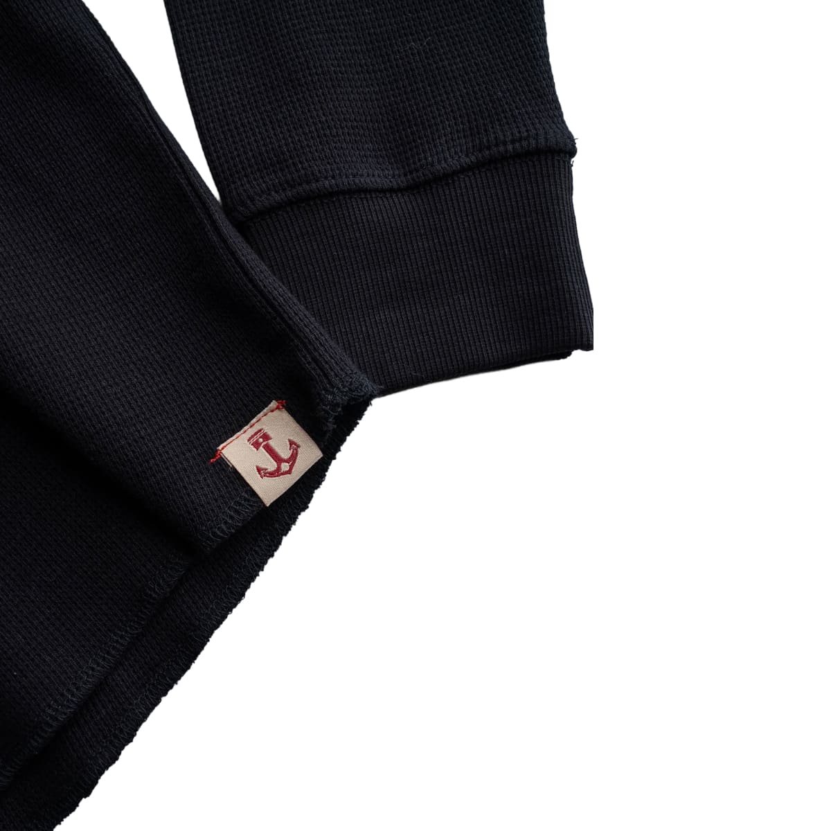Close-up of a black Iron and Resin Cotton Crewe Neck with a small beige brand tag featuring a red anchor logo, shown against a white background.