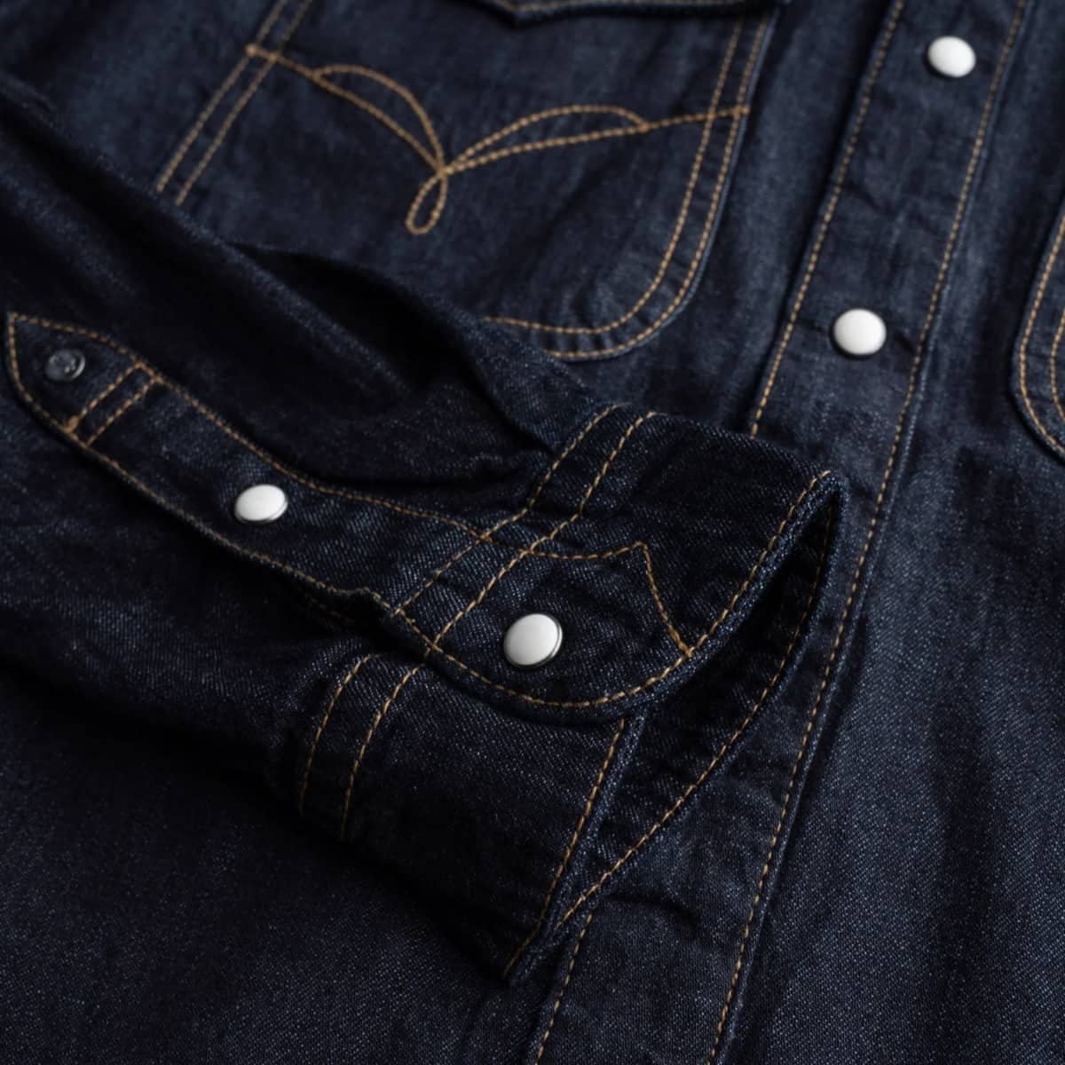 Close-up of a dark indigo denim shirt jacket showing the chest pocket with contrast yellow stitching, a buttoned cuff, and white snap buttons.