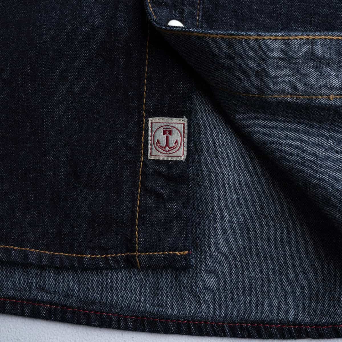 Close-up of dark indigo denim fabric with contrast tan stitching and a small square brand patch featuring an anchor logo.