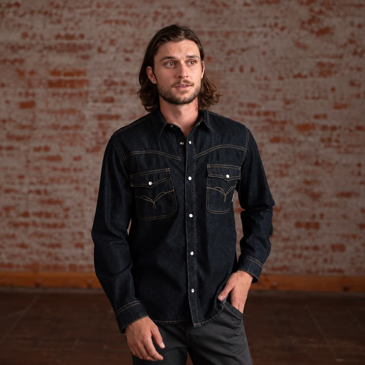 A man wearing a dark indigo denim western-style shirt with contrast stitching and button details, standing in front of a rustic brick wall.