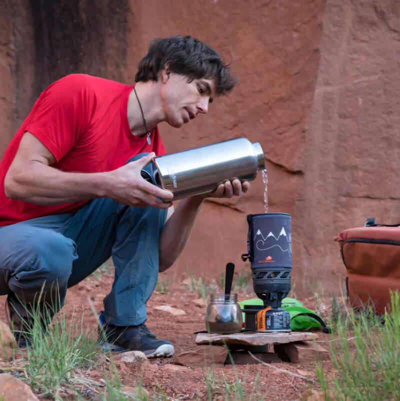 Person in a red shirt pouring liquid from a thermos into a camping coffee maker outdoors.