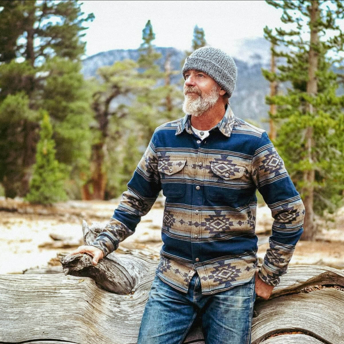 Man in a patterned shirt and jeans standing outdoors with trees and mountains in the background