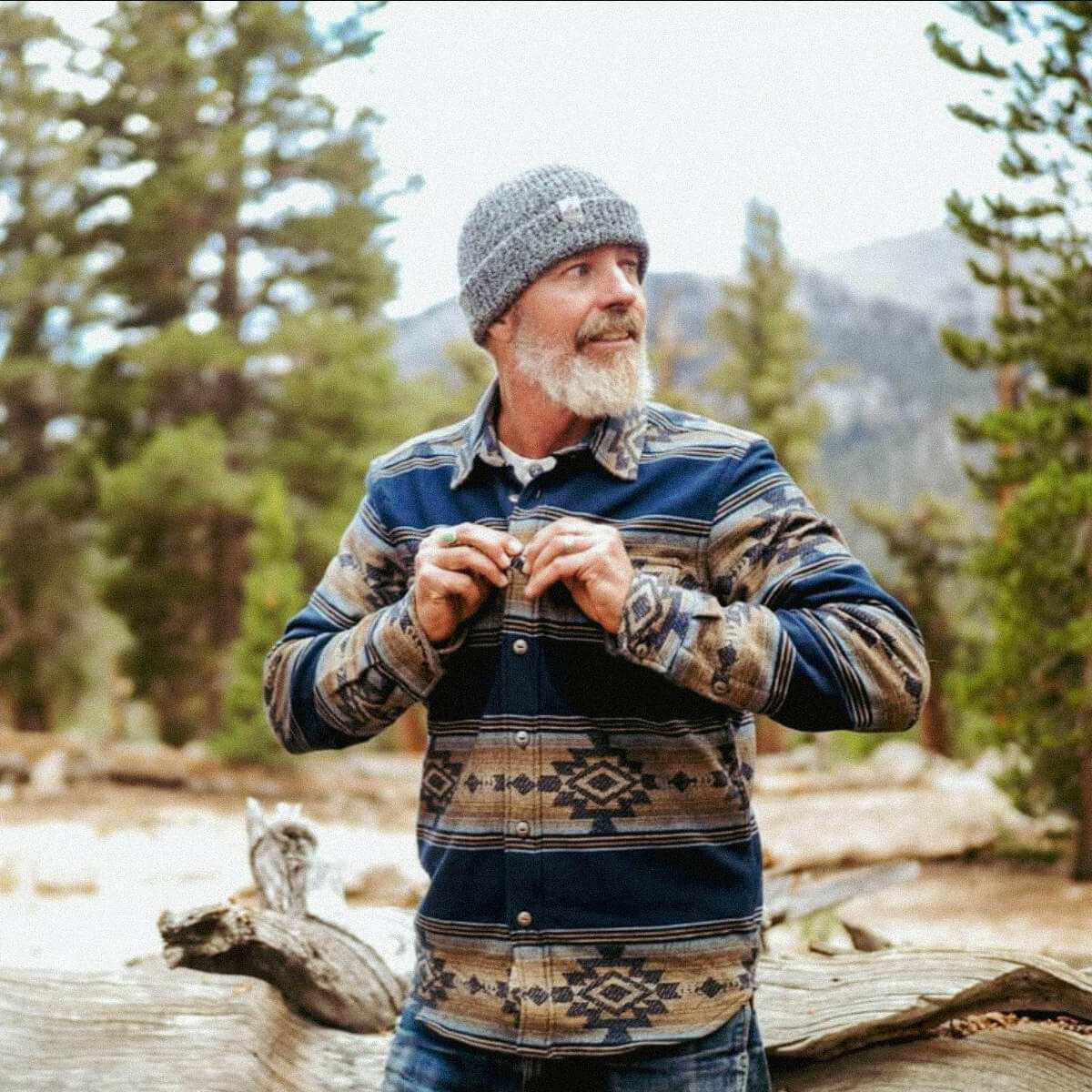 Man in a patterned jacket and knit hat standing in a forest with mountains in the background