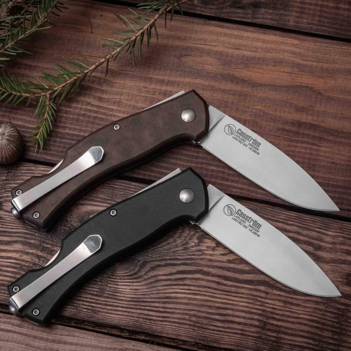 Two folding knives on a wooden surface with a branch and pine cones.