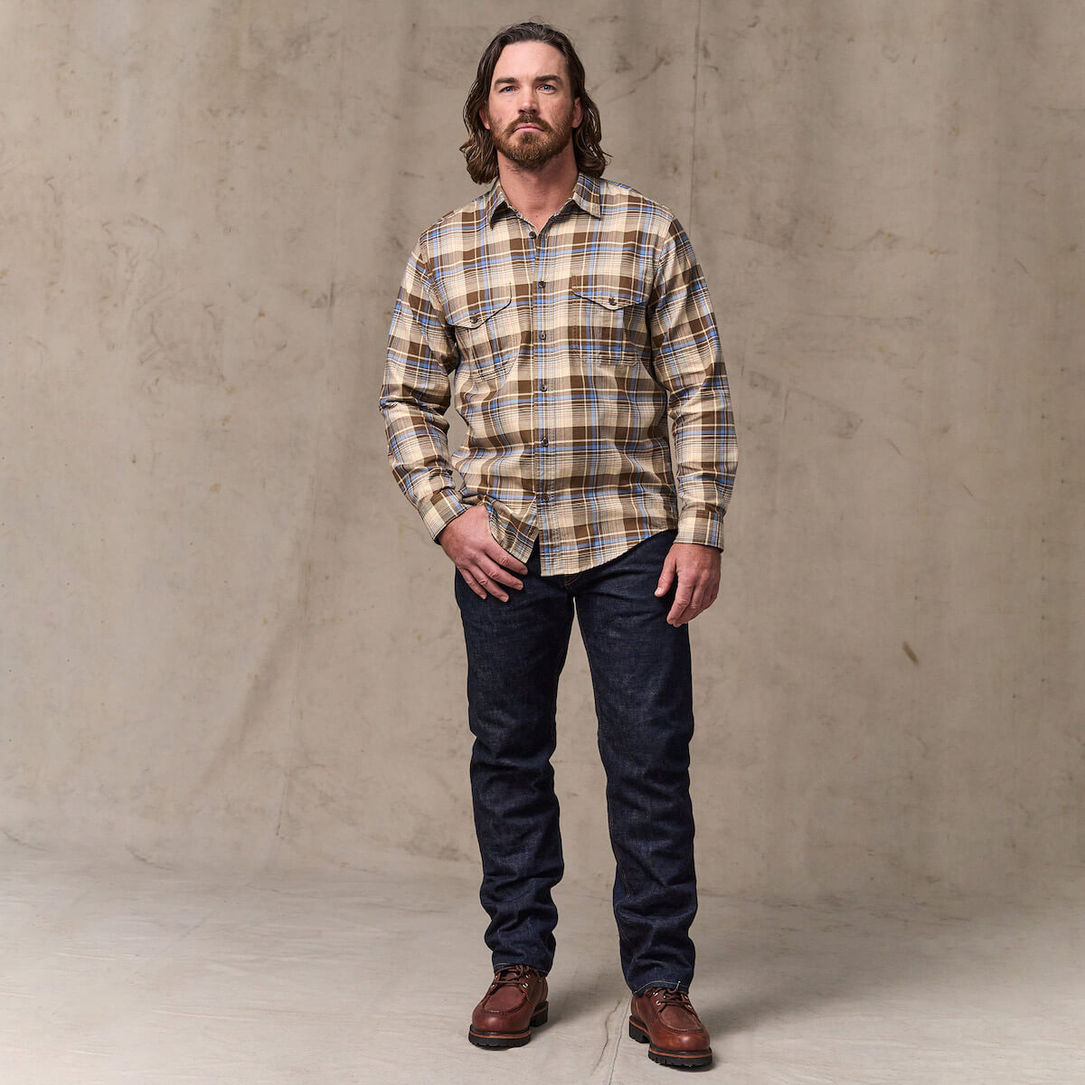 Man wearing a plaid shirt and dark jeans standing against a plain background