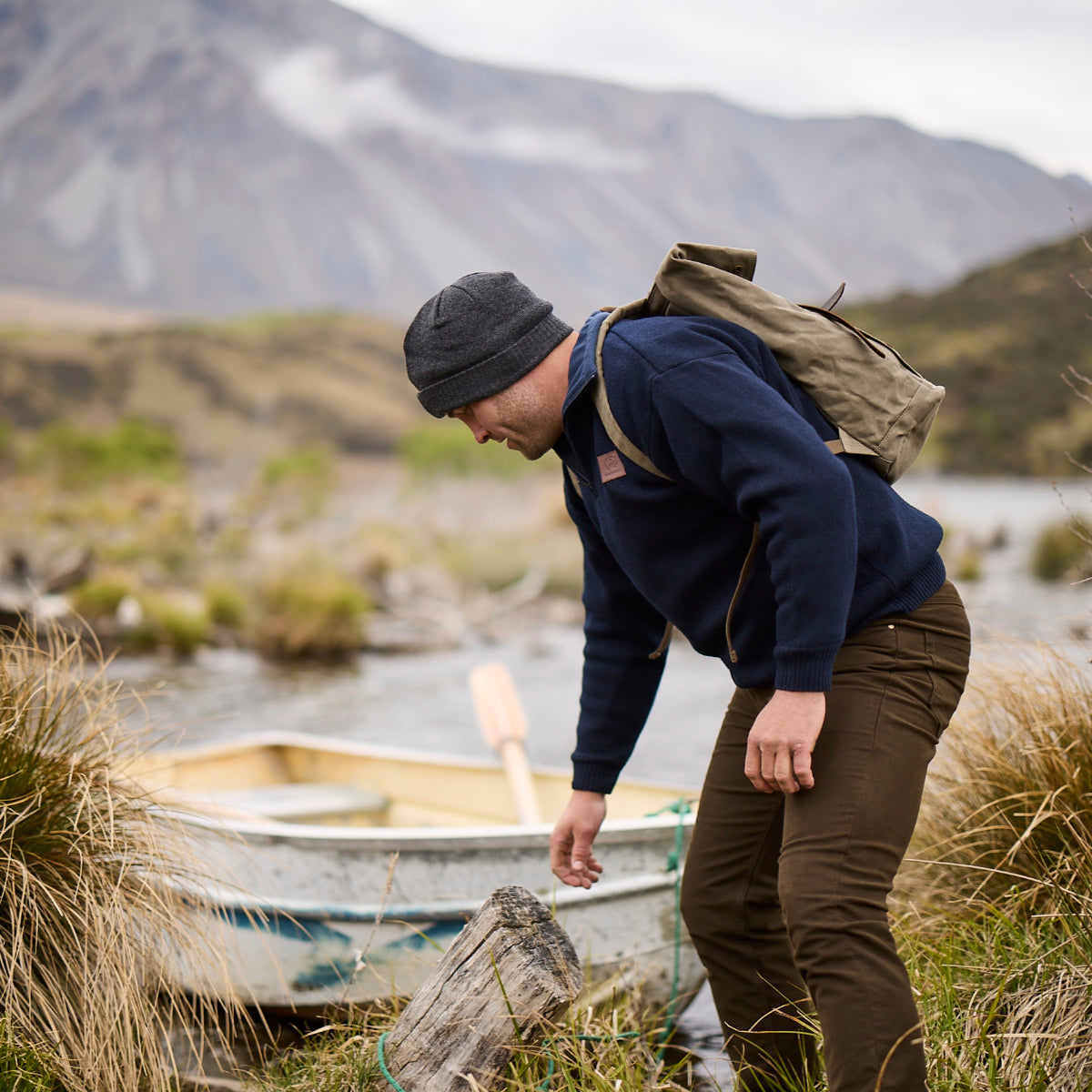 A man in a navy fleece sweater, brown  Swanndri pants, and a dark beanie bends down near a small boat on a riverbank, with mountains and grassy hills in the background. He is wearing a tan canvas backpack.