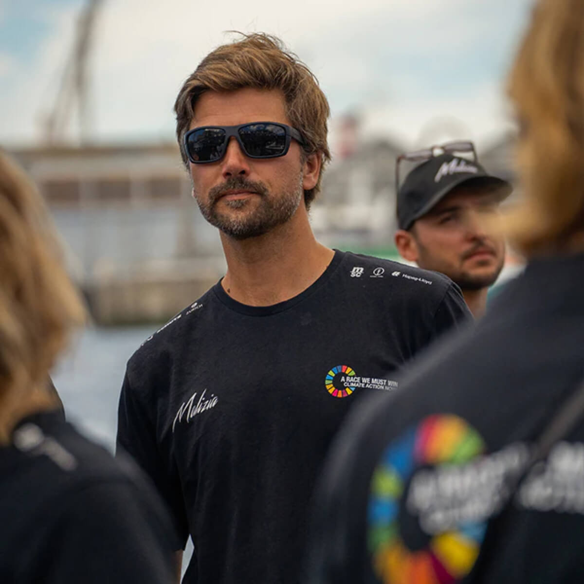 Man wearing sunglasses and a black shirt with logos, standing outdoors near water.