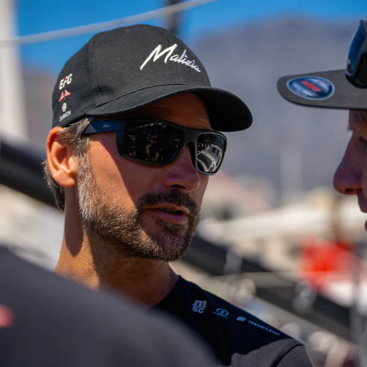 Man wearing a black cap with visible branding, sunglasses, and a blurred background