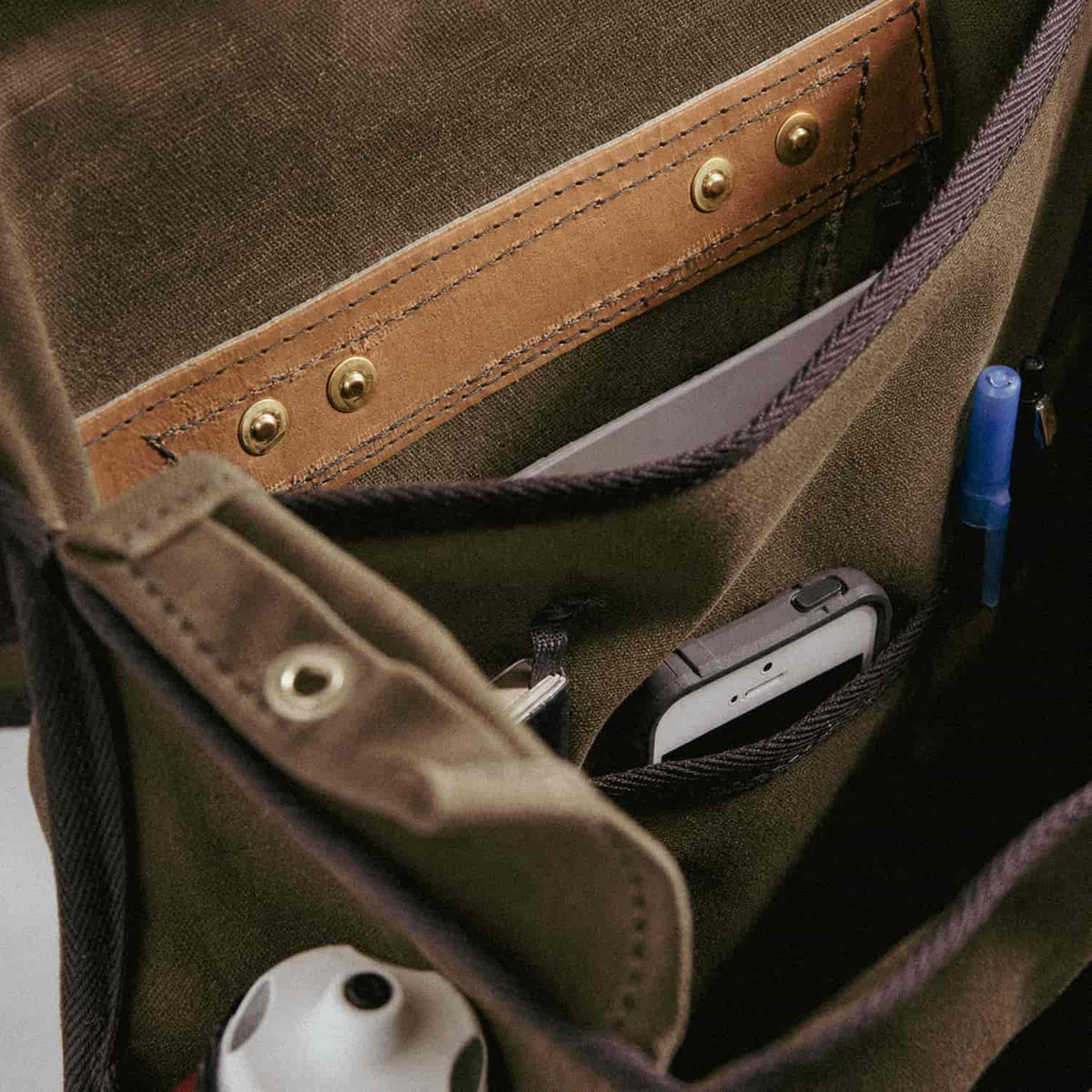 Close-up of a Frost River organiser pouch with multiple compartments, showing a tan leather trim, metal rivets, and stored items including a white device, a blue pen, and a small accessory.