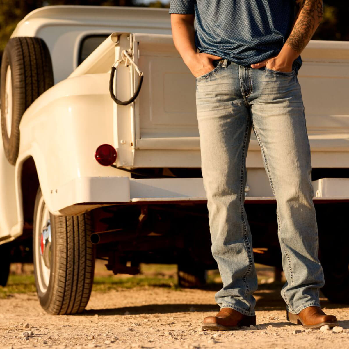 Person wearing blue jeans standing next to a white truck on a dirt road.