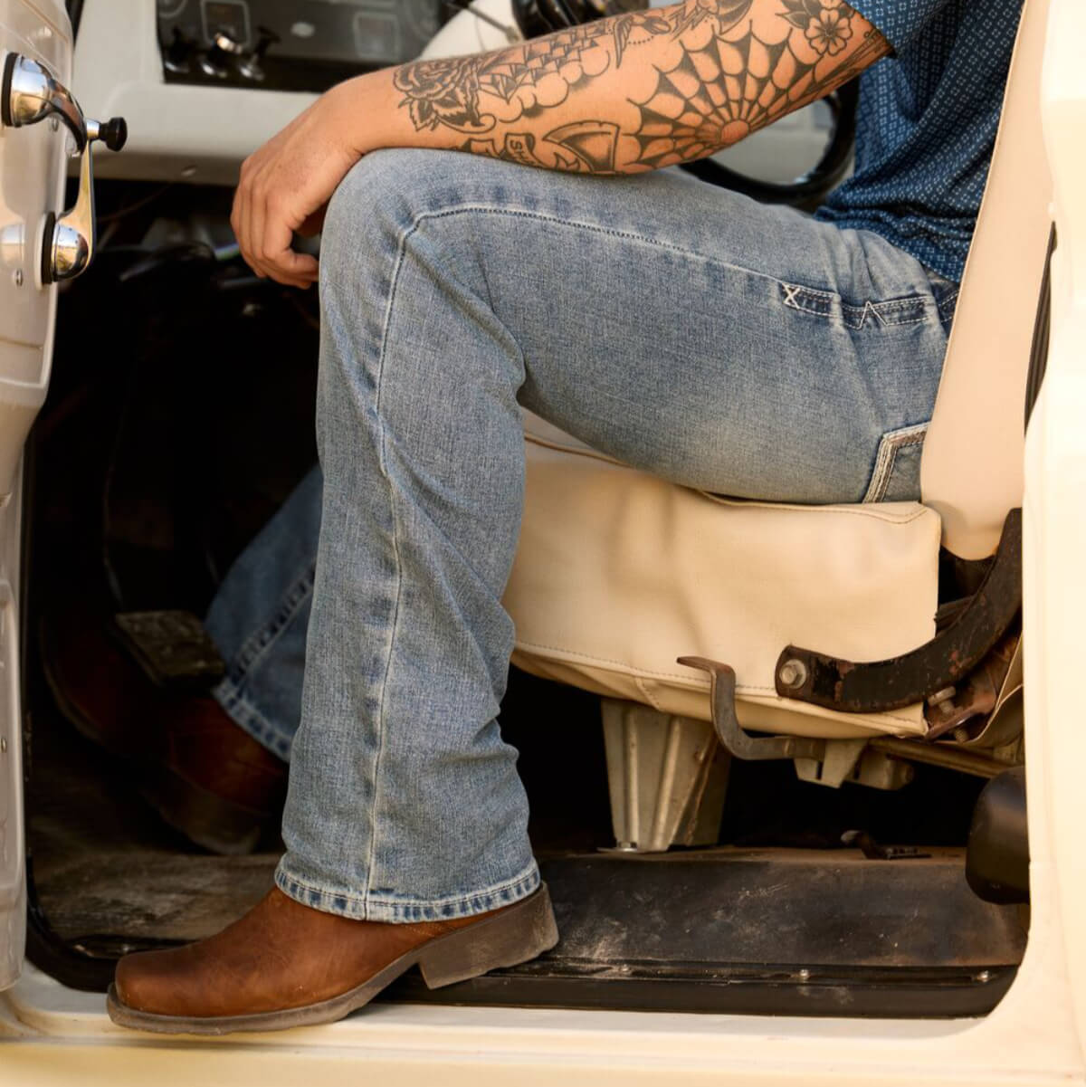 Person sitting inside a vehicle wearing blue jeans and brown boots, with a tattooed arm visible.