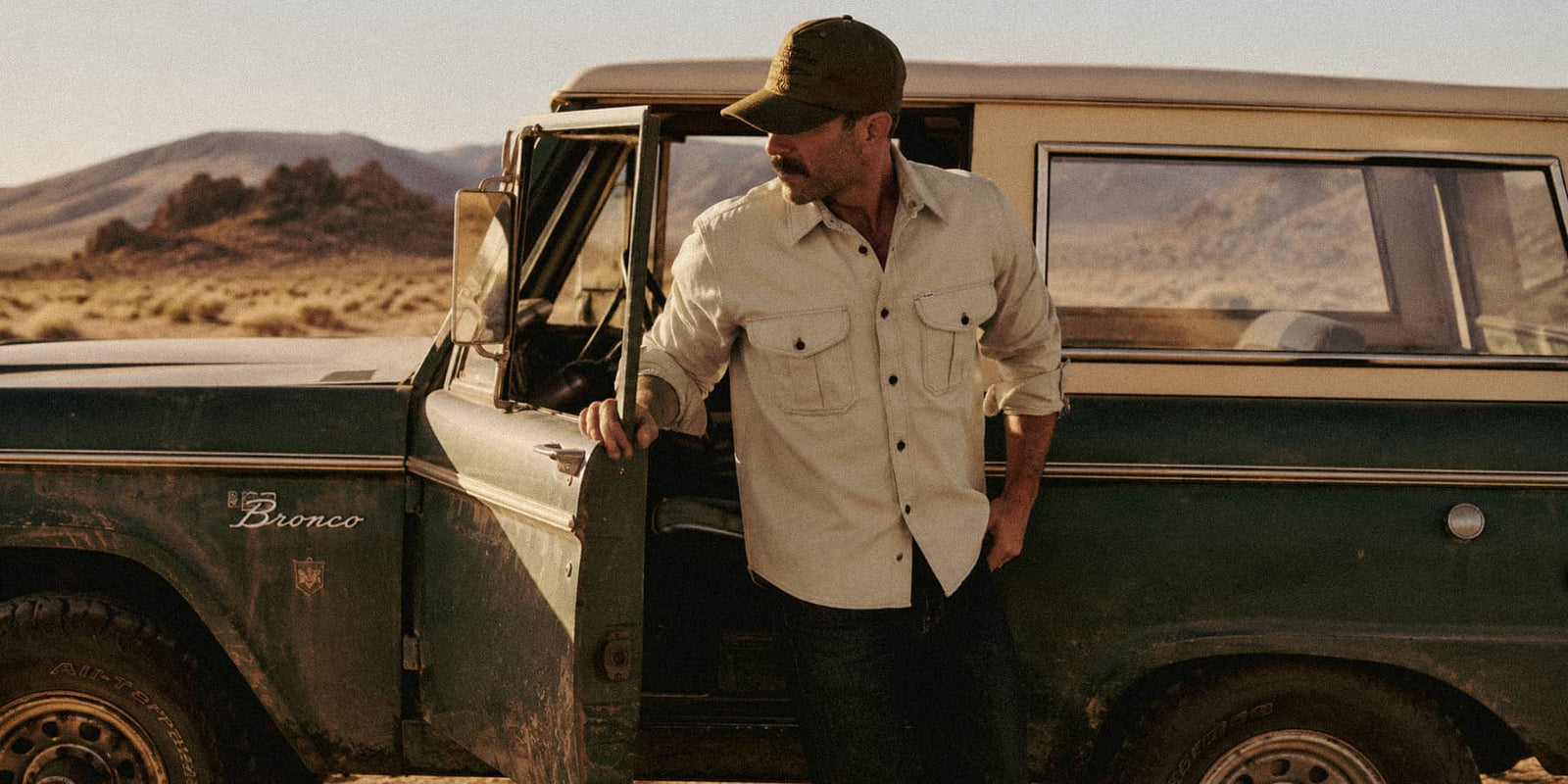 A man wearing a Filson guide shirt and a green cap leans against the open door of a vintage green and beige station wagon in a desert landscape.