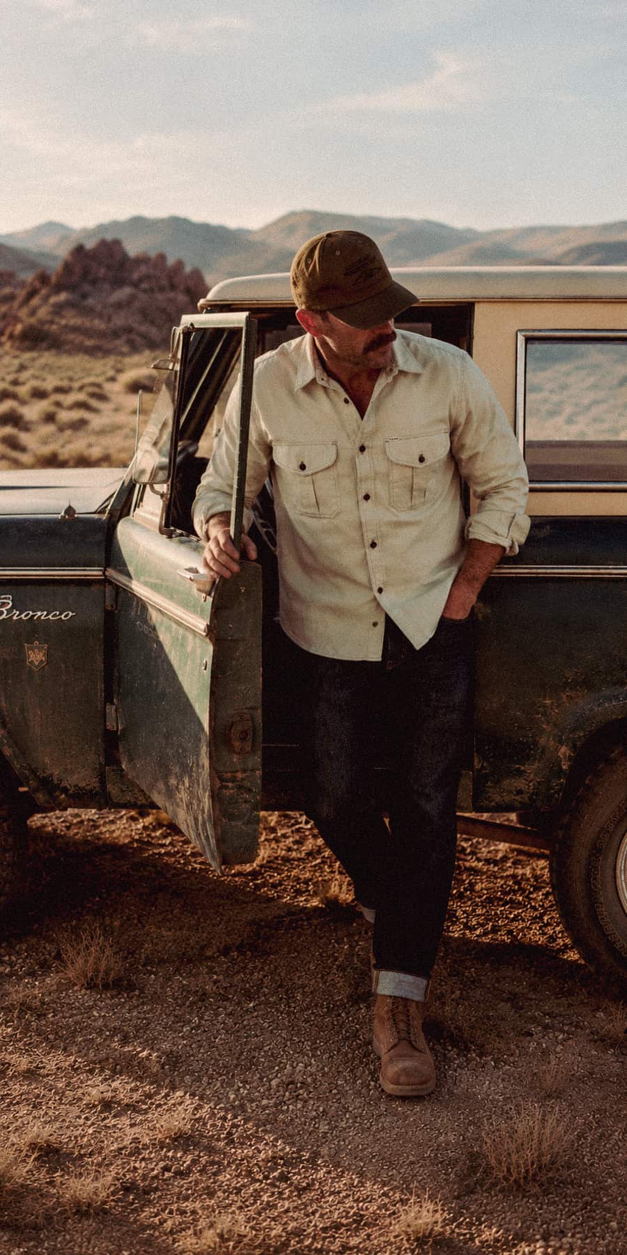 A man wearing a Filson button-up shirt, dark jeans, brown boots, and a brown cap leans against the open door of a vintage off-road truck in a desert landscape.
