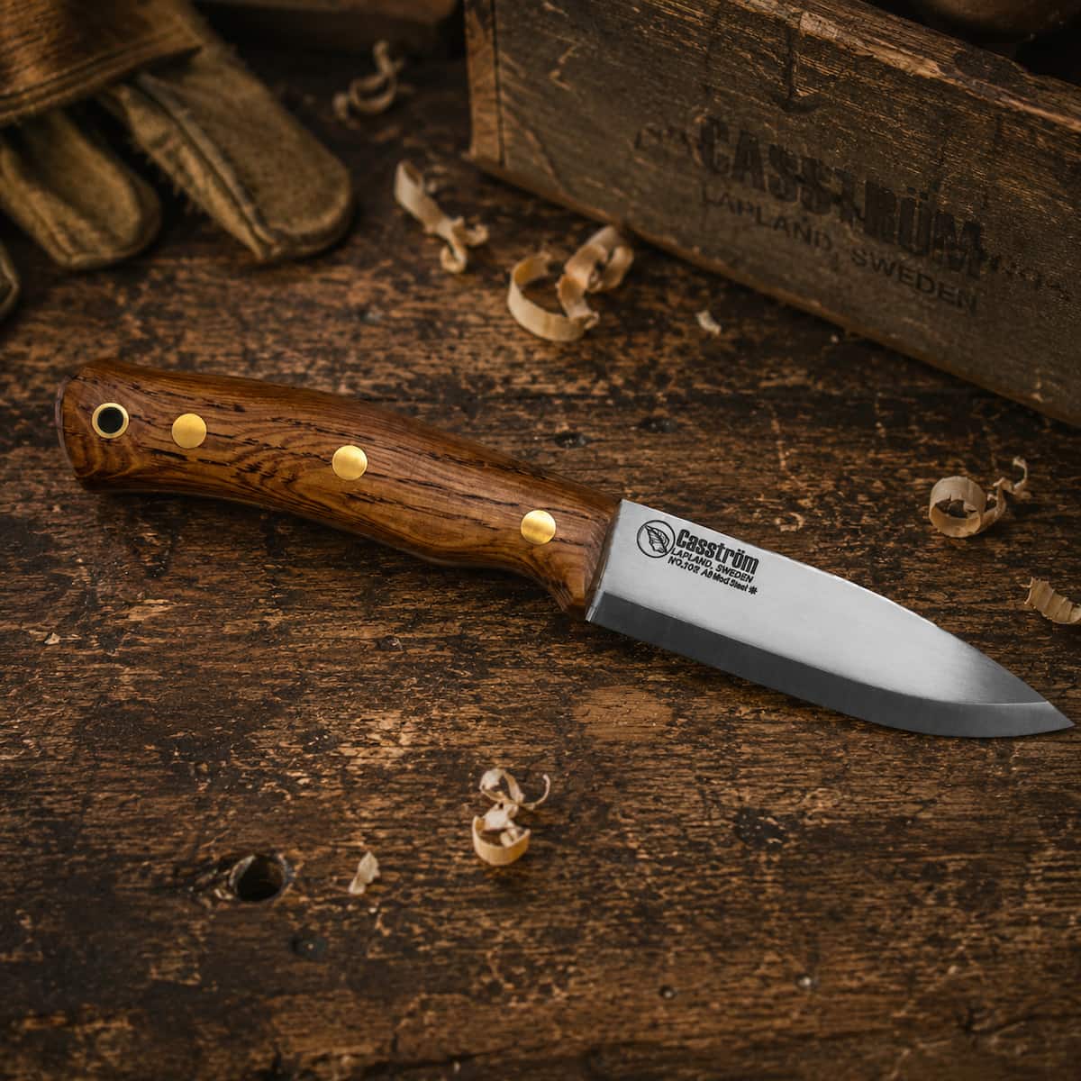 A small fixed-blade knife with a polished steel blade and an oiled wood handle lies on a rustic wooden workbench. The handle features brass rivets and a visible 'Casstrom' brand logo on the blade.