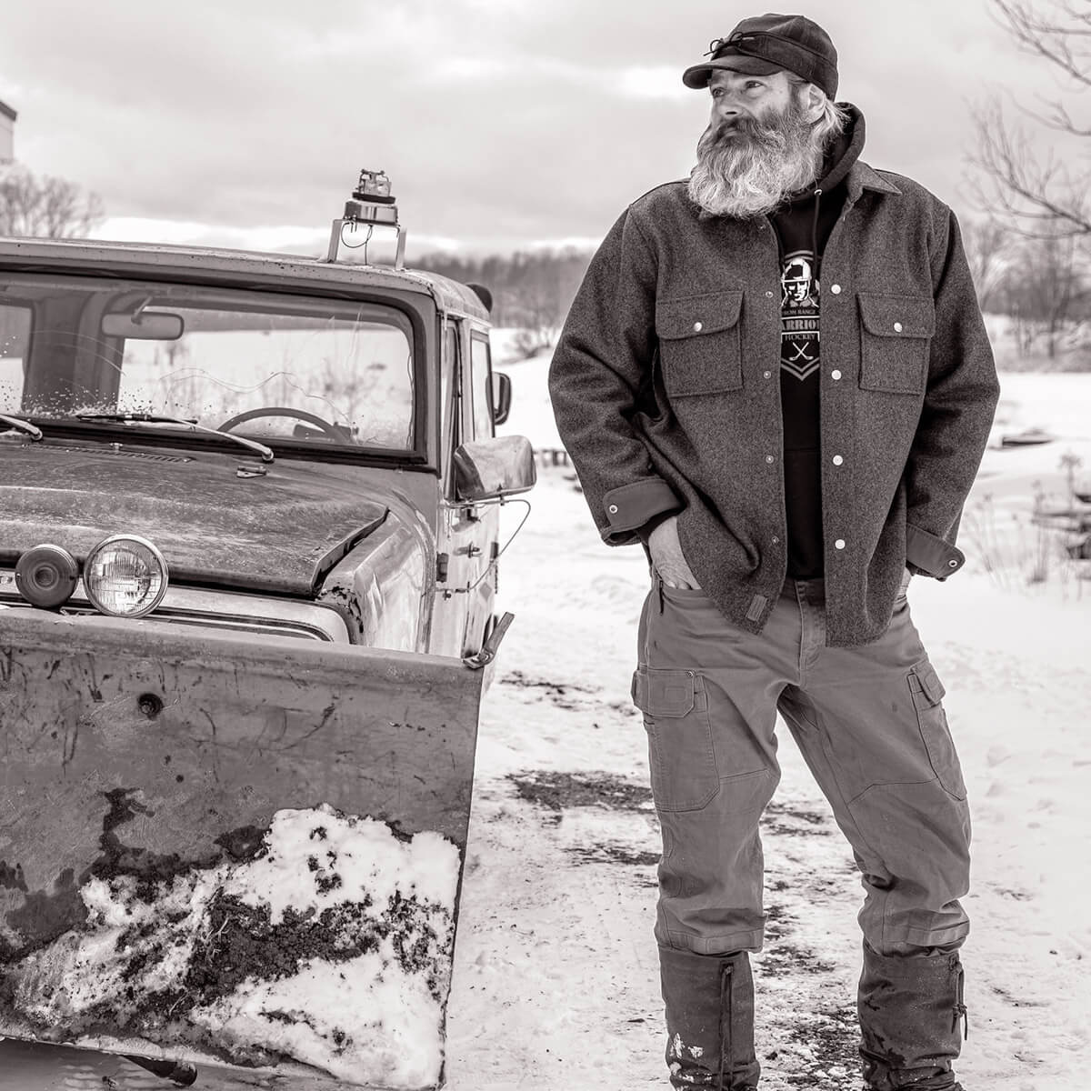 Man with a beard and cap standing next to an old truck in a snowy landscape