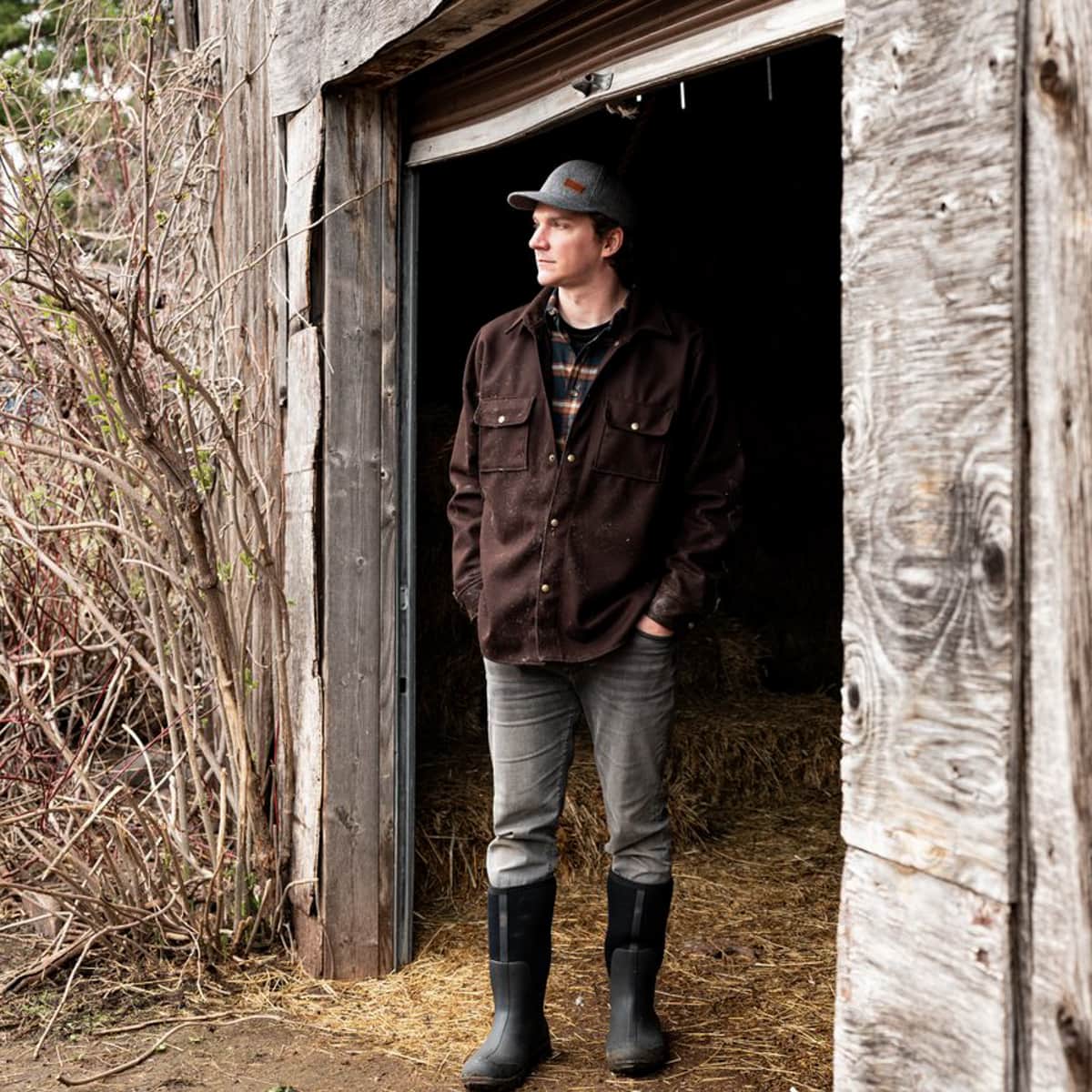 A man wearing a Stormy Kromer Wool Overshirt, gray jeans, black rubber boots, and a gray cap stands in the doorway of a rustic wooden barn.