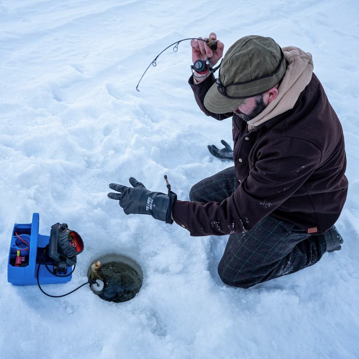 A man kneeling in the snow while ice fishing, holding a fishing rod above an open hole. A blue electric ice fishing machine sits to the left, and he wears a brown jacket, olive green hat, and black gloves in a snowy outdoor setting.
