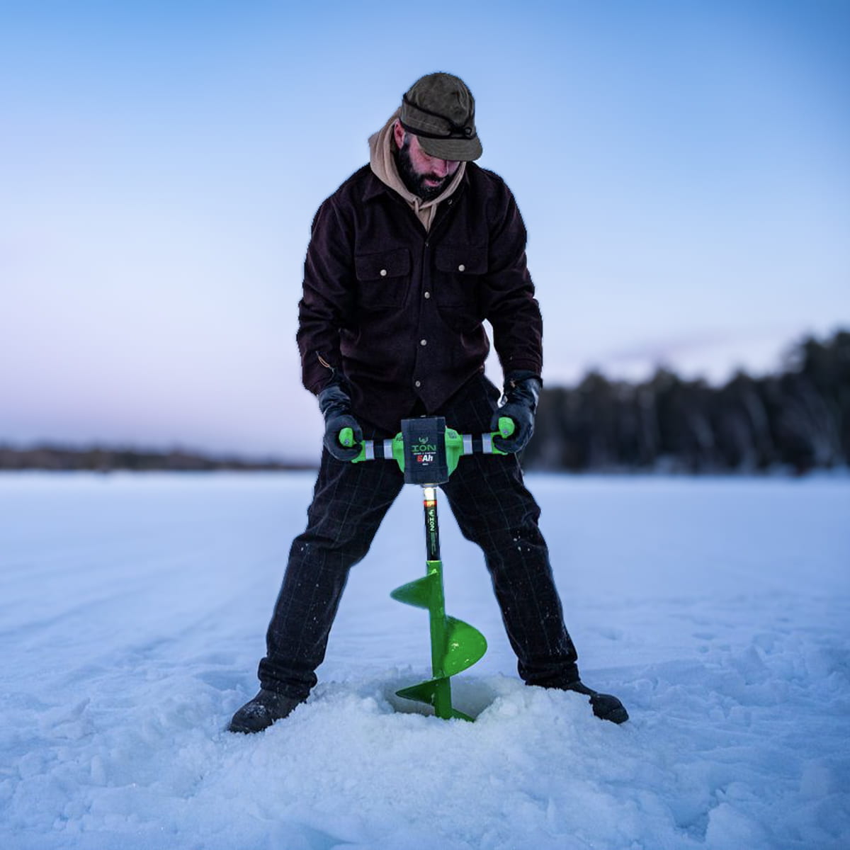 A man wearing winter gear stands on a frozen lake using a green and black ice auger to drill a hole in the snow. The background shows a snowy landscape and a soft blue twilight sky.