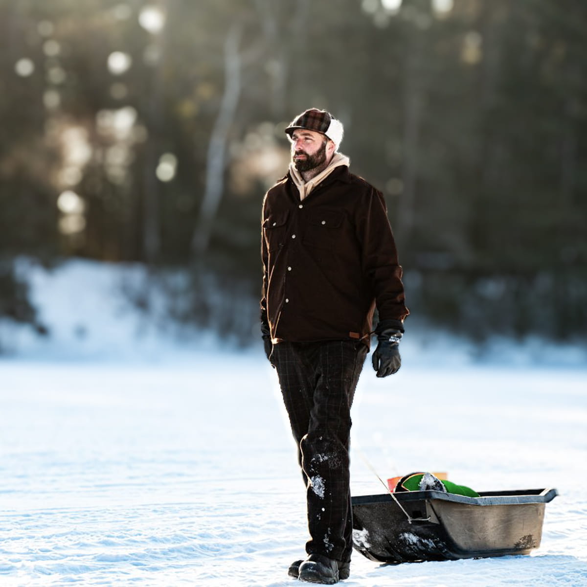 A bearded man wearing a brown jacket, dark pants, and a cap stands on a snowy outdoor surface next to a metal sled.