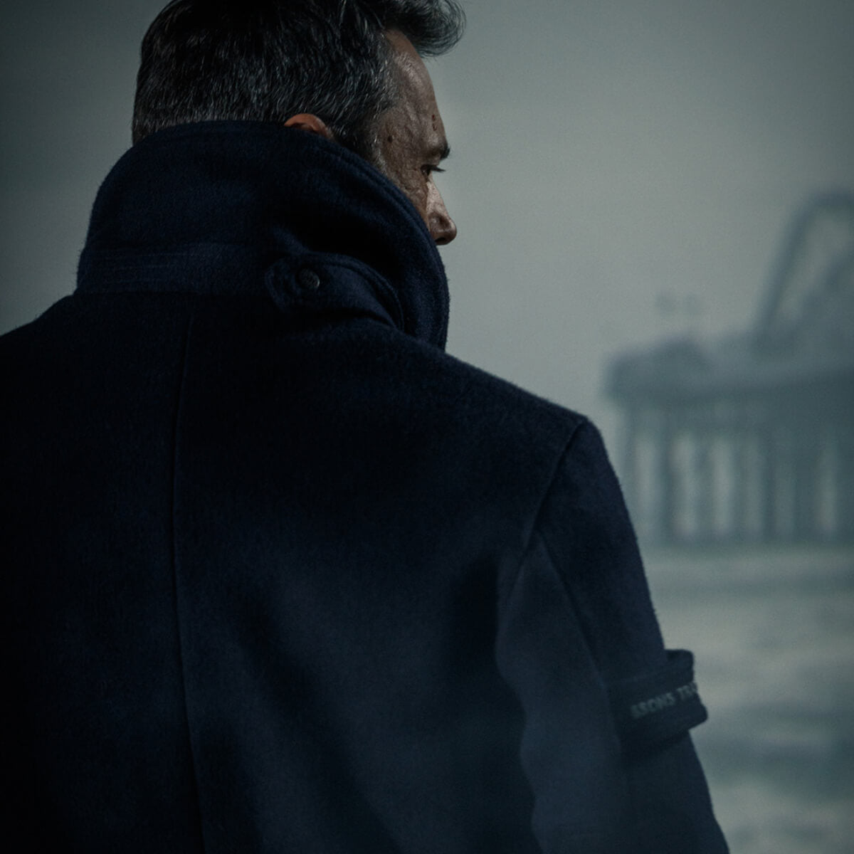 Man in a dark coat looking out over water with a misty pier in the background