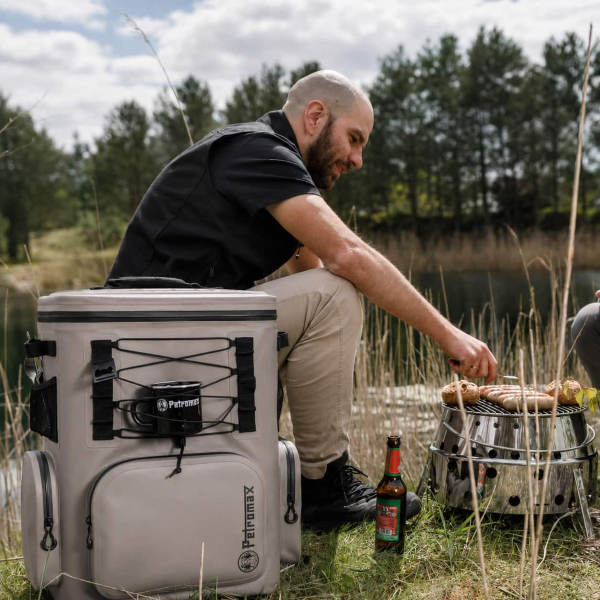 A man outdoors by a lake and trees is kneeling beside a large beige PerforMax cooler while grilling sausages on a small metal stove. A bottle is on the ground nearby.