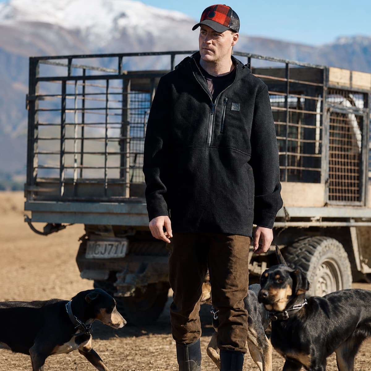 A man wearing a black fleece pullover and brown pants stands outdoors in front of a truck with dogs at his side, with mountains in the background.